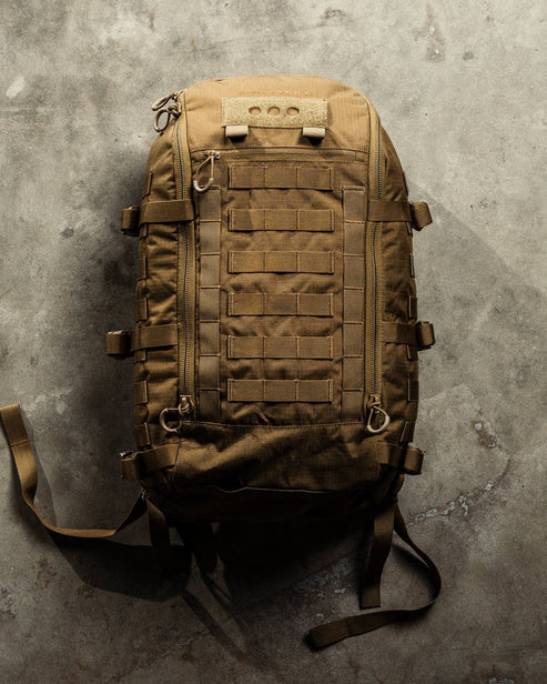 Ein brauner EBERLESTOCK Mission Assault Pack mit modularem Aufbau, mehreren Gurten, Schlaufen, Befestigungsgurten und Sicherungsschnallen liegt auf einem Betonboden.
