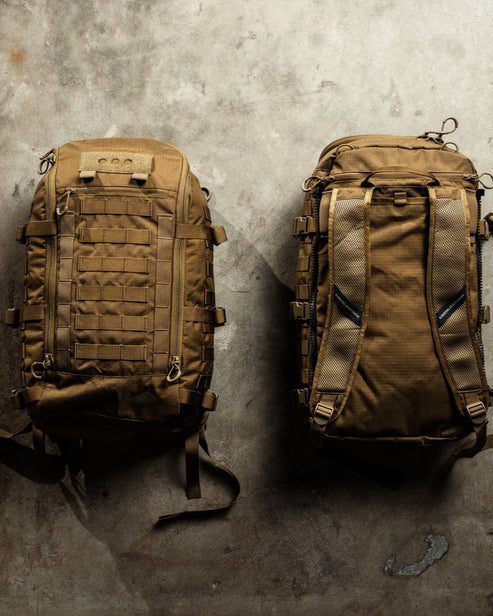 Zwei hellbraune EBERLESTOCK Mission Assault Packs liegen auf dem Beton - einer ist nach vorne gerichtet und zeigt mehrere Gurte und Taschen, der andere zeigt gepolsterte Schultergurte und eine Netzrückseite, die den modularen Aufbau eines Einsatzrucksacks verdeutlicht.