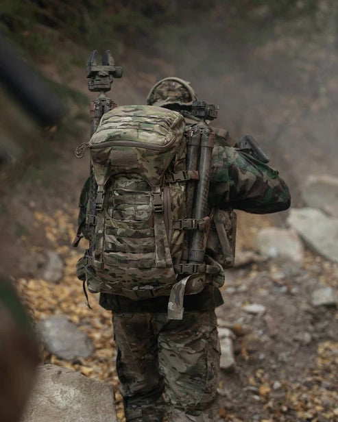 Ein Soldat in Camouflage geht mit dem Rücken zur Kamera auf einem felsigen Waldweg und trägt einen EBERLESTOCK Mission Assault Pack. Umgeben von Bäumen und herabgefallenen Blättern ist das modulare Design des Rucksacks sichtbar.
