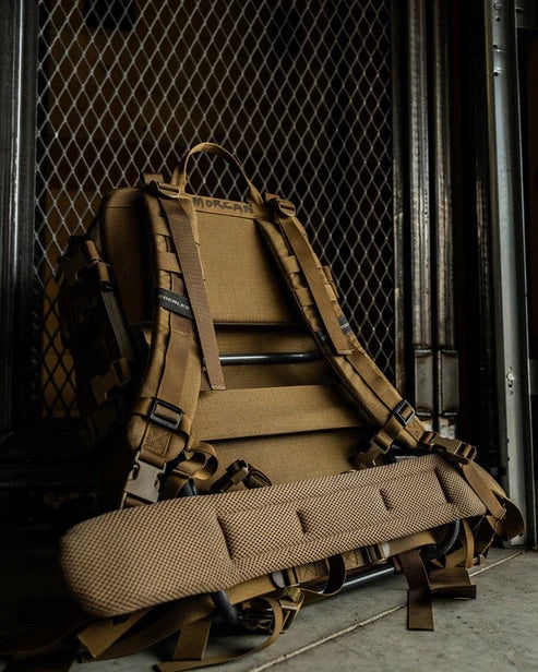 Der EBERLESTOCK Mission EMOD Frame, ein hellbrauner taktischer Rucksack mit gepolsterten Trägern und Hüftgurt, steht im Innenbereich an einem Metallgitterzaun und zeigt seine robuste Bauweise, die zahlreichen Befestigungspunkte und den modularen Rahmen für flexible taktische Ausrüstung.
