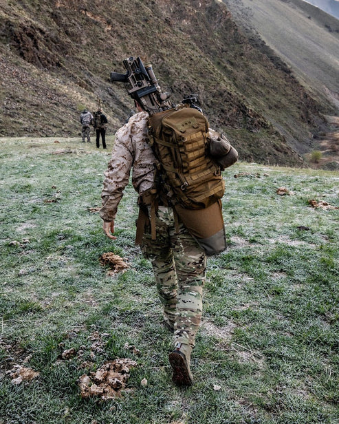 Ein Soldat in Camouflage mit einem modularen EBERLESTOCK Mission EMOD Frame Rucksackgestell wandert mit einem Gewehr einen grasbewachsenen, felsigen Hang hinauf. Zwei weitere Soldaten in taktischer Ausrüstung stehen vor ihm auf dem zerklüfteten, bergigen Hang.