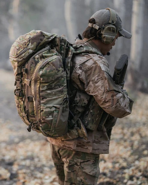 Eine Person in Tarnkleidung und mit Mütze trägt einen EBERLESTOCK Mission EMOD Frame Rucksack und hält ein Gewehr in einem Wald mit herabgefallenen Blättern - vorbereitet mit taktischer Ausrüstung für die freie Natur.