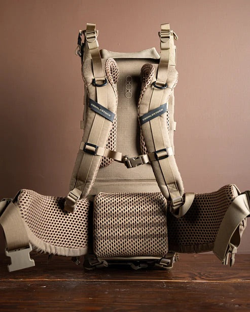 Ein hellbrauner EBERLESTOCK Mainframe II Rucksack mit ergonomischem Tragesystem, gepolsterten Schultergurten, Netzrückenstütze und breitem Hüftgurt steht aufrecht auf Holz vor einem schlichten braunen Hintergrund.