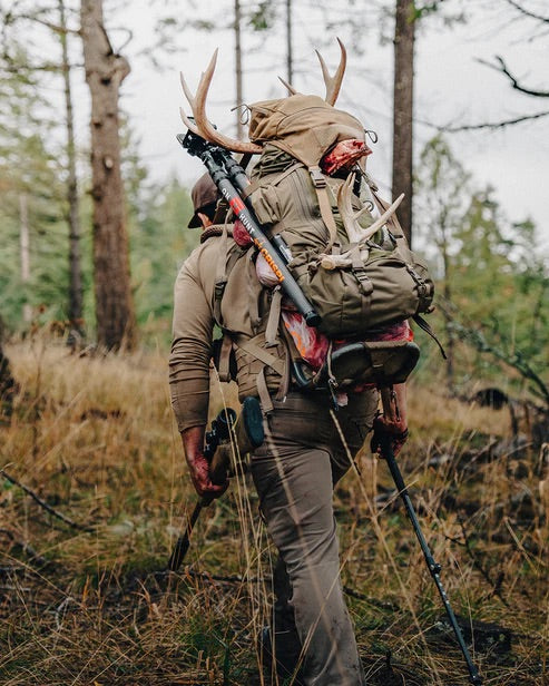 Ein Abenteurer, ausgerüstet mit dem EBERLESTOCK Mainframe II, wandert durch den Wald, trägt Hirschgeweih und Fleisch mit Trekkingstöcken in der Hand - ein idealer Begleiter für jede Jagdexpedition zwischen hohen Bäumen und trockenem Gras.