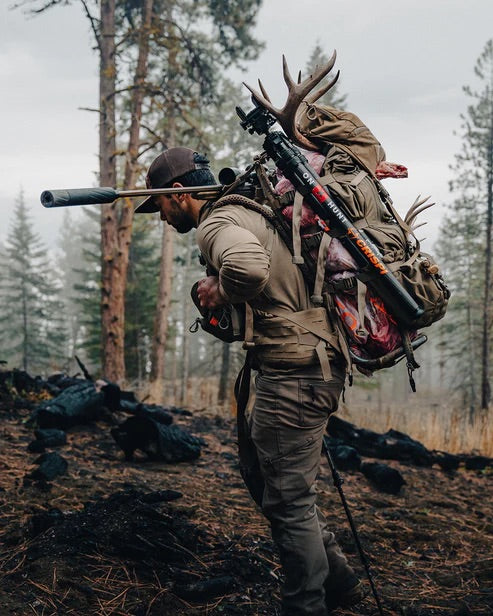 Ein Jäger in Outdoor-Ausrüstung wandert durch einen nebligen Wald und trägt ein Geweih, ein Gewehr und Vorräte mit der EBERLESTOCK Mainframe II. Bäume und Nebel unterstreichen die raue Atmosphäre in der Wildnis.