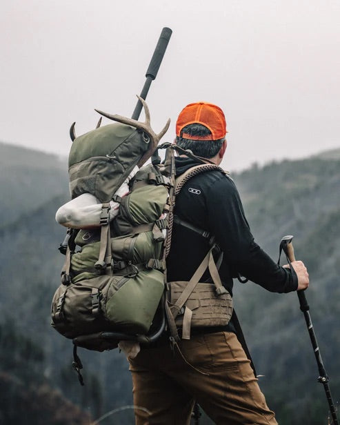 Mit orangefarbener Mütze und dem EBERLESTOCK Mainframe II Rucksack mit aufgesetztem Geweih steht ein Trekker mit Trekkingstöcken auf einem nebligen Bergpfad, überblickt den Wald und ist bereit für die Jagd.