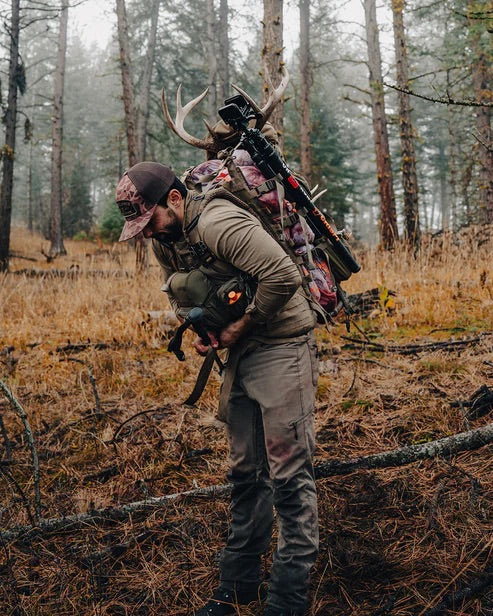 Eine Person in Outdoor-Ausrüstung und mit Mütze steht in einem nebligen Wald mit einem EBERLESTOCK Eberlestock Mainframe II, der ein Geweih und einen Compoundbogen trägt, und blickt nach unten, während sie die Jagdausrüstung hält.