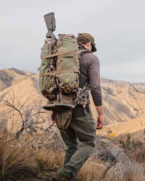 In Outdoor-Ausrüstung und mit einem EBERLESTOCK Mainframe II Rucksack, an dem ein Gewehr befestigt ist, läuft eine Person durch trockenes Gras - ideal für die Jagd inmitten von Hügeln und Bergen im Hintergrund.