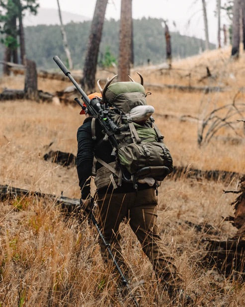 Für die Jagd mit Outdoor-Ausrüstung ausgestattet, trägt eine Person ein Gewehr und einen EBERLESTOCK Mainframe II Rucksack mit aufgesetztem Geweih, während sie durch einen trockenen, grasbewachsenen Wald geht.