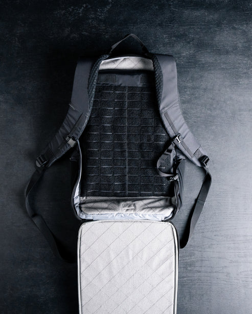 Ein schwarzer Rucksack wird geöffnet und flach auf eine dunkle Oberfläche gelegt. Zum Vorschein kommt ein gitterartig gemustertes Innenleben mit dem EBERLESTOCK Fade Blocker Sleeve Tablet als Teil der gesteppten Polsterung im Hauptfach.