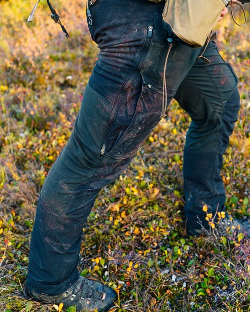 Eine Person in schwarzer, schmutzbefleckter EBERLESTOCK Deadwood Pant - Gunmetal und Wanderstiefeln steht auf einer grasbewachsenen Herbstwiese. Diese Hose ist ein praktischer, wasserabweisender Begleiter für Outdoor-Abenteuer inmitten von buntem Laub und kleinen Pflanzen.