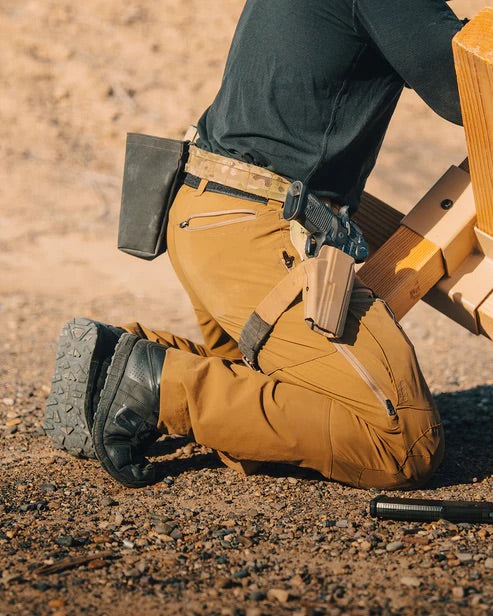 Eine Person in EBERLESTOCK Deadwood Pant - Gunmetal, schwarzen Stiefeln und einem schwarzen langärmeligen Hemd kniet auf dem Kies neben einer Holzkonstruktion und trägt einen Gürtel mit Taschen und eine Pistole im Holster.