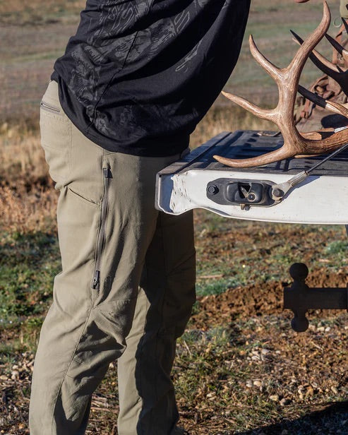 Eine Person, die eine EBERLESTOCK Camas Pro Pant - Gunmetal und ein schwarzes Hemd trägt, steht an der Heckklappe eines Lastwagens und hält mit einer Hand ein Geweih auf Erde und Gras im Freien.