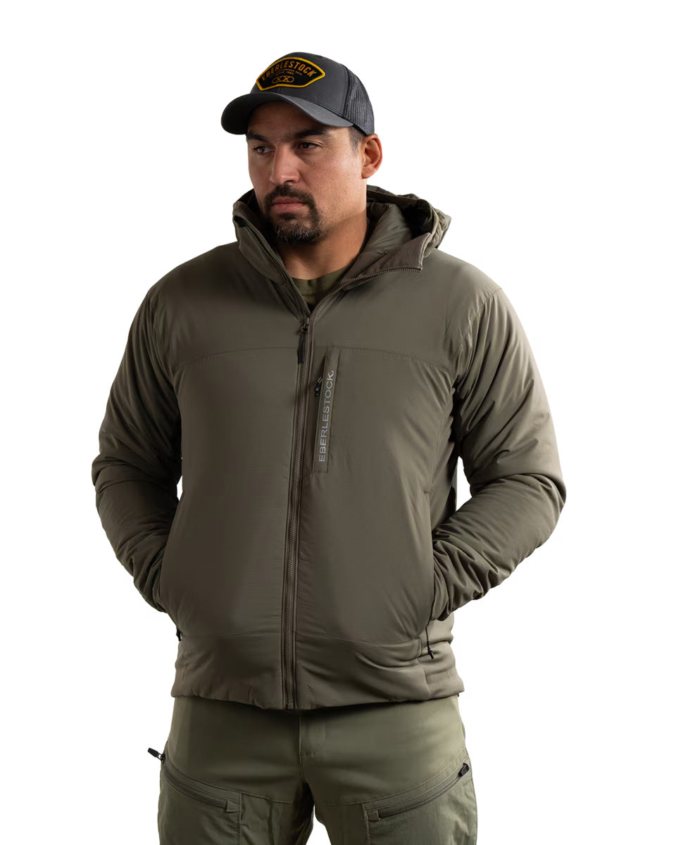 Ein Mann trägt die EBERLESTOCK Alturas Hooded Jacket - Ranger Green, eine olivgrüne Hose und eine schwarze Baseballkappe und steht mit den Händen in den Taschen vor einem schlichten weißen Hintergrund.