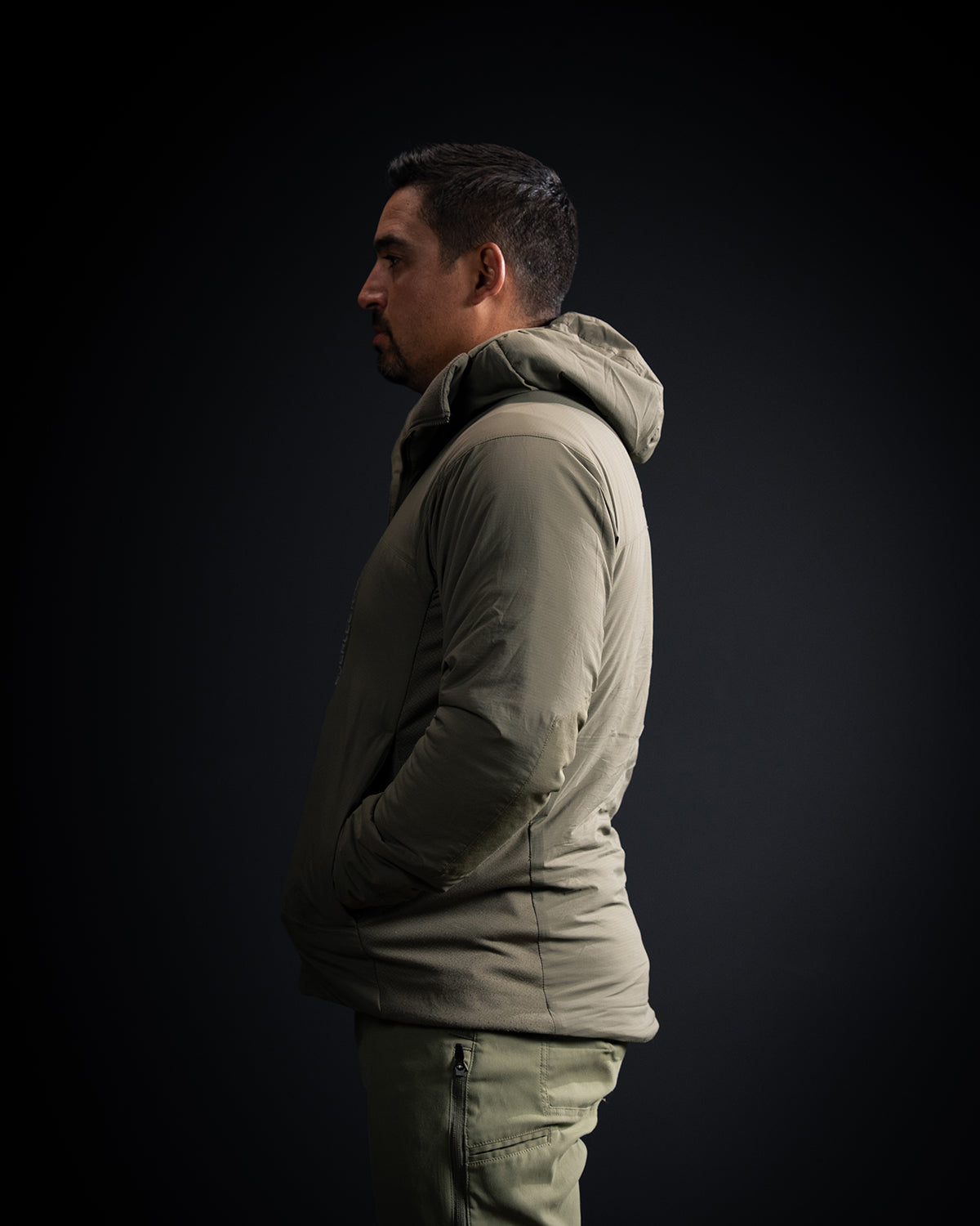 Ein Mann mit kurzen dunklen Haaren steht im Profil vor einem dunklen Hintergrund, die Hände in den Taschen, und trägt die EBERLESTOCK Alturas Hooded Jacket - Ranger Green und eine passende Hose.
