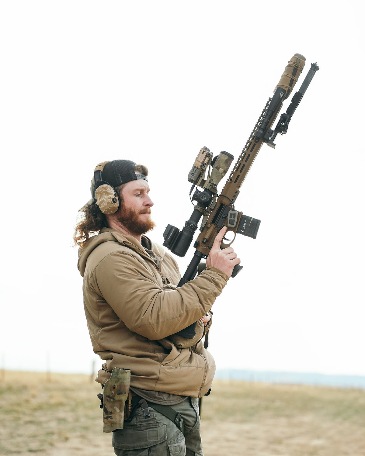 Ein Mann mit langen Haaren und Bart, der eine EBERLESTOCK Alturas Kapuzenjacke in Ranger Green, einen Gehörschutz und eine Mütze trägt, steht im Freien und hält ein großes Gewehr mit Zielfernrohr nach oben gerichtet, während er eine Pistole im Holster an seiner Hüfte trägt.