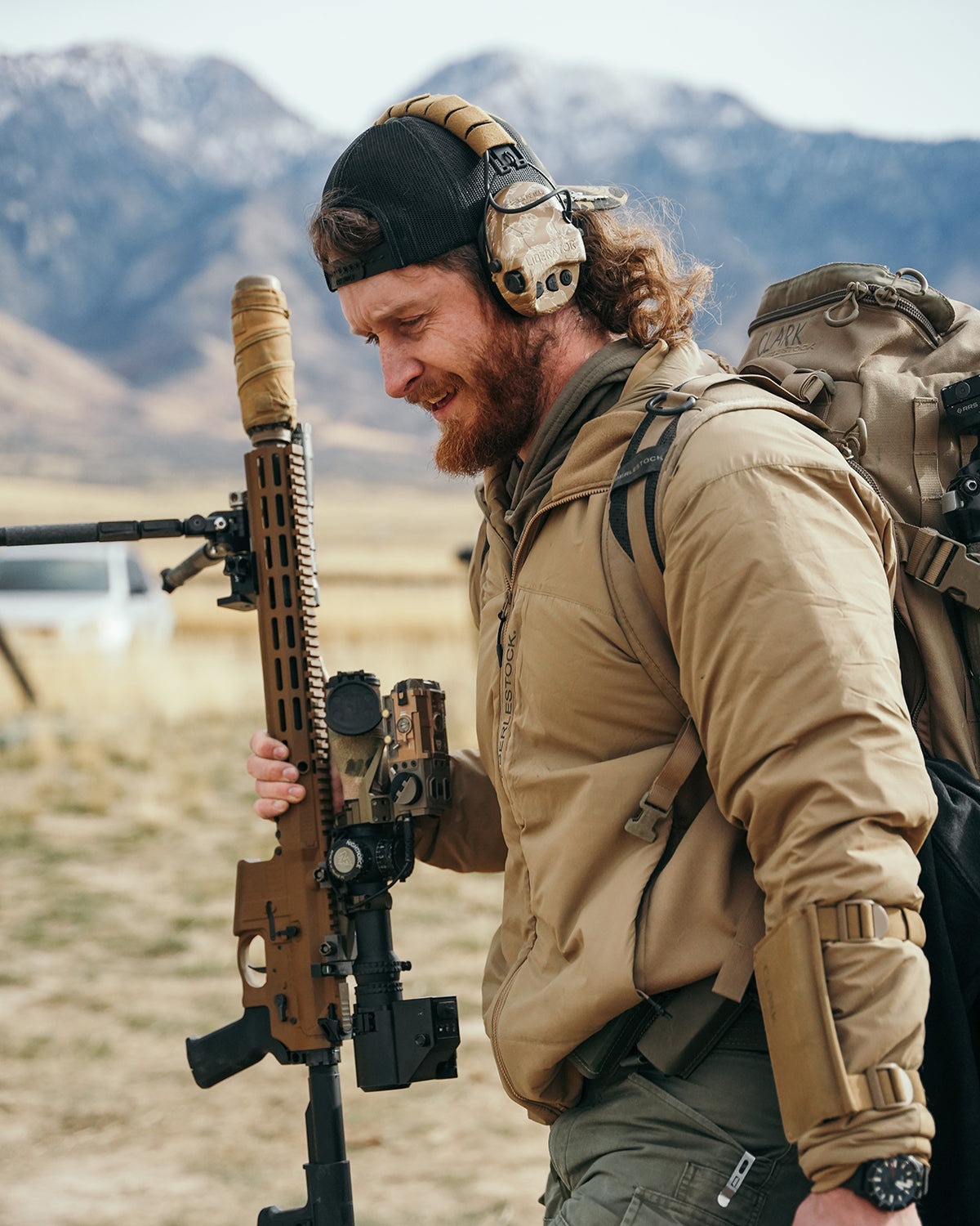 Ein bärtiger Mann mit Gehörschutz, schwarzer Mütze und einer EBERLESTOCK Alturas Hooded Jacket in Ranger Green hält ein hellbraunes Gewehr mit Schalldämpfer auf einem Outdoor-Schießstand mit den Bergen im Hintergrund.