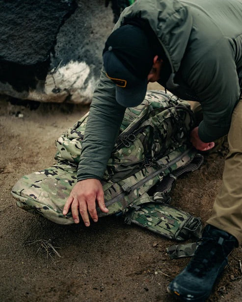 Eine Person in Outdoor-Kleidung und Stiefeln hockt auf sandigem Boden neben einem großen Felsen und passt einen EBERLESTOCK Eberlestock Spectre Rucksack mit Tarnmuster an.
