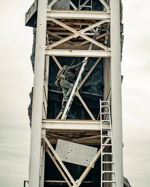 Bekleidet mit Outdoor-Kleidung und einem Eberlestock Spectre von EBERLESTOCK klettert eine Person unter bedecktem Himmel auf eine Metallleiter an einem hohen weißen Industriegebäude, in dessen Nähe sich eine weitere Leiter befindet.