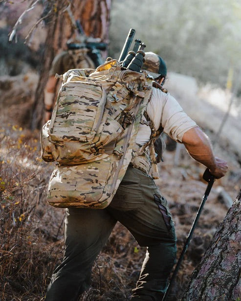 Eine Person in Camouflage, die den ultraleichten EBERLESTOCK Eberlestock Spectre Rucksack trägt, wandert mit Trekkingstöcken bergauf durch einen Wald. Ein anderer, ähnlich gekleideter Wanderer ist vor ihm zu sehen.