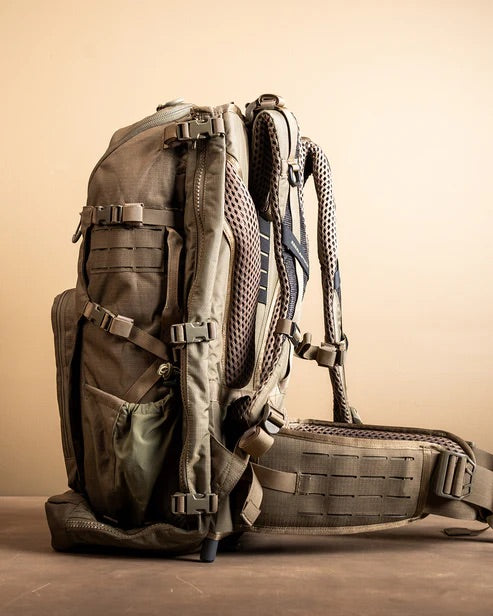 Der taktische Rucksack EBERLESTOCK Spectre in Hellbraun und Olivgrün mit mehreren Gurten, Taschen und Netzpolsterung steht aufrecht auf einer braunen Oberfläche mit beigem Hintergrund.
