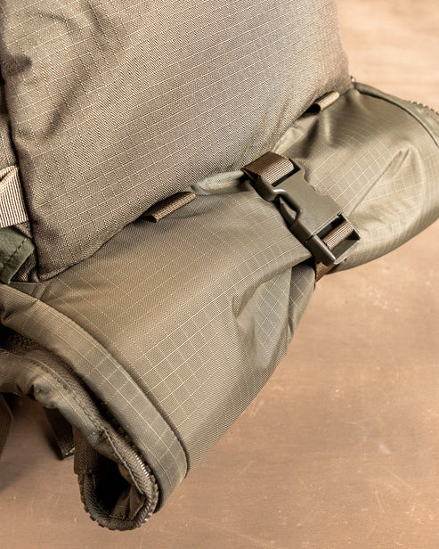 Nahaufnahme des EBERLESTOCK Spectre Rucksacks in khakigrün mit Gittermuster, mit einem aufgerollten, durch eine schwarze Kunststoffschnalle gesicherten Teil, der auf einer braunen Oberfläche ruht.