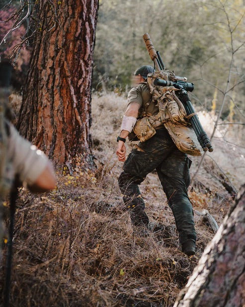 Eine Person in Tarnhose mit einem großen Gewehr und einem EBERLESTOCK Eberlestock Spectre Rucksack erklimmt einen bewaldeten Hügel, während im Vordergrund eine weitere, teilweise sichtbare Person erscheint.