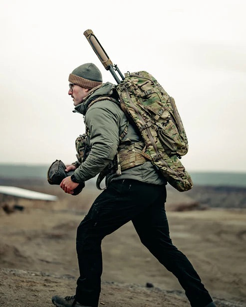 Ein Mann in Outdoor-Kleidung und mit Mütze trägt den EBERLESTOCK Spectre, einen großen, ultraleichten Jagdrucksack in Tarnfarben mit Ausrüstung und Gewehr, während er bei bedecktem Himmel bergauf durch unwegsames Gelände wandert.