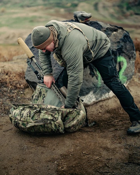 Eine Person in Outdoor-Ausrüstung beugt sich über eine EBERLESTOCK Eberlestock Spectre Tarntasche, während sie mit einem Gewehr hantiert, mit einer Graslandschaft und einem großen Felsen im Hintergrund.