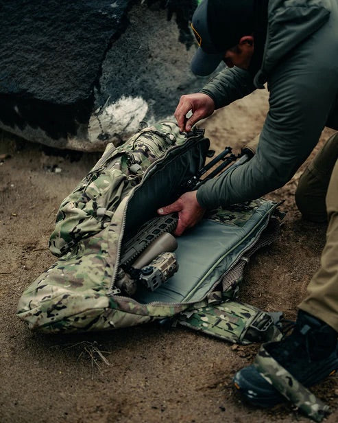 Eine Person in Outdoor-Ausrüstung kniet im Sand und öffnet den Reißverschluss eines EBERLESTOCK Eberlestock Spectre Rucksacks. Zum Vorschein kommen ein Gewehr und ein Stativ - ein Zeichen dafür, dass sie sich auf die Jagd oder das Schießen im Freien vorbereitet.