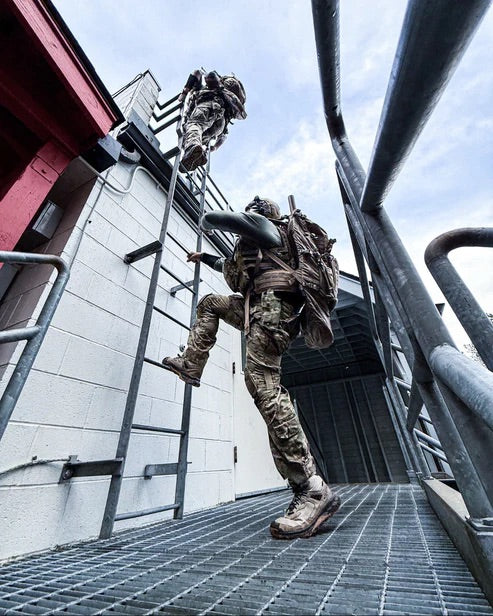 Zwei Soldaten in Tarnuniformen und Helmen, die jeweils einen Eberlestock Spectre von EBERLESTOCK tragen, klettern eine Metallleiter hinauf, die an einem Gebäude befestigt ist, das von unten auf einer vergitterten Plattform zu sehen ist, eine höher gelegene mit dem Himmel darüber.