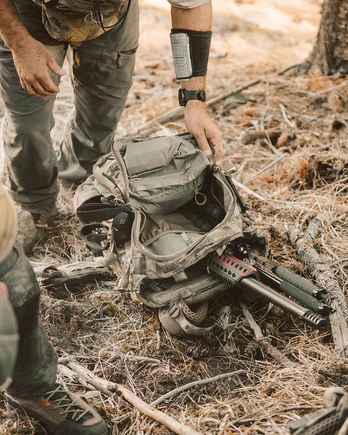 Im Freien öffnet eine Person einen EBERLESTOCK Eberlestock Spectre Rucksack auf dem Boden und legt ein Gewehr und eine Ausrüstung inmitten von trockenem Wald mit verstreuten Stöcken und Tannennadeln frei.