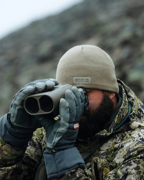 Mit Camouflage, grauen Handschuhen und der EBERLESTOCK North Fork Merino Beanie trägt eine Person ein Fernglas in felsigem Gelände - eine ideale Outdoor-Kopfbedeckung.