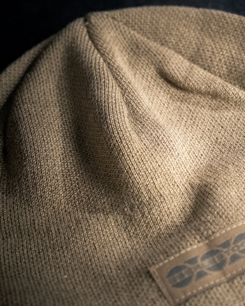 Nahaufnahme der Eberlestock North Fork Merino Beanie von EBERLESTOCK mit ihrem strukturierten beigen Merinowollstrick und einem teilweise sichtbaren rechteckigen Etikett mit dunklem Muster in der unteren rechten Ecke.