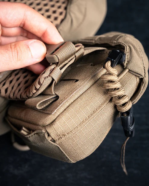 Eine Nahaufnahme zeigt eine Hand, die einen Clip an einem hellbraunen taktischen Rucksack von EBERLESTOCK einstellt. Er ist mit Mesh-Polsterung, Reißverschlüssen und einer aufgerollten Kordel ausgestattet, daneben befindet sich eine Eberlestock Mag-Free Rangefinder Pouch.