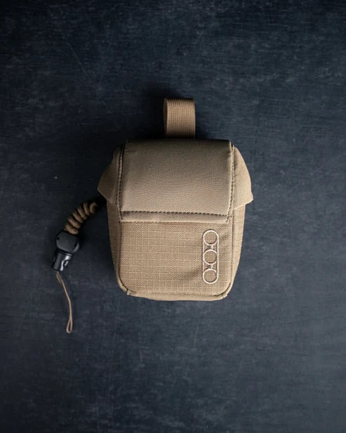 Eine kompakte, braune EBERLESTOCK Mag-Free Rangefinder Pouch mit Klappdeckel, Schlaufengriff und drei runden Metallringen ruht auf einer dunklen Oberfläche; eine aufgewickelte Kordel mit schwarzem Clip ist an dieser Tasche befestigt.