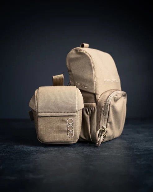 Zwei beigefarbene EBERLESTOCK Mag-Free Rangefinder Pouches ruhen auf einer dunklen Oberfläche mit schwarzem Hintergrund. Die größere Tasche hat mehrere Fächer, während die kleinere Fronttasche ein einfaches Logo mit vier Kreisen zeigt.