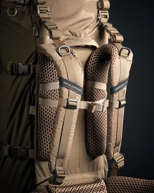 Nahaufnahme des braunen EBERLESTOCK Brute 6500 Rucksacks mit gepolsterten, netzgefütterten Schultergurten und robusten Schnallen, der für den Einsatz im Freien oder bei taktischen Einsätzen konzipiert ist, vor einem dunklen Hintergrund.