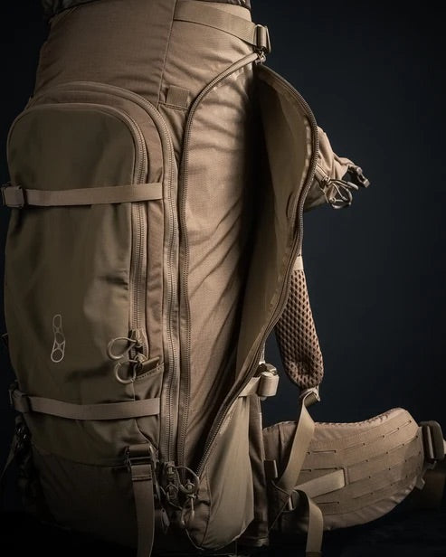 Ein hellbrauner EBERLESTOCK Brute 6500 Expeditionsrucksack mit mehreren Reißverschlussfächern und gepolsterten Gurten steht aufrecht vor einem dunklen Hintergrund. Eine Seitentasche mit offenem Reißverschluss offenbart ihr Netzfutter.