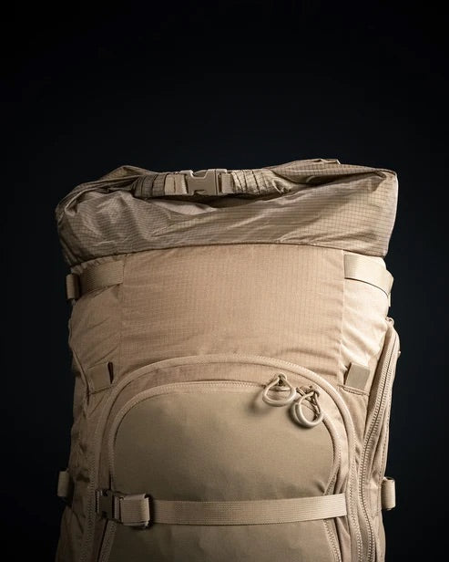 Ein großer brauner EBERLESTOCK Brute 6500 taktischer Rucksack mit Rollverschluss, Schnallenriemen, Vordertasche mit Reißverschluss und Karabinerhaken wird vor einem dunklen Hintergrund fotografiert.