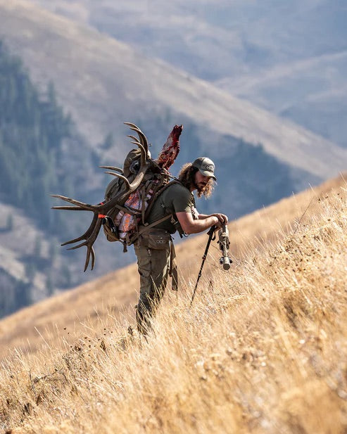 Ein Jäger wandert bergauf durch trockenes Gras und trägt Geweih, Fleisch und Ausrüstung in seinem EBERLESTOCK Brute 6500 Rucksack. Mit Wanderstöcken als Stütze geht er auf ferne Berge und ein bewaldetes Tal zu.