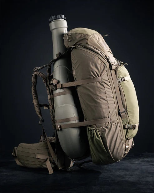 Der EBERLESTOCK Brute 4500, ein großer hellbrauner und grüner Militärrucksack mit gepolsterten Trägern und mehreren Fächern, ist vor einem dunklen Hintergrund mit einem langen zylindrischen Behälter abgebildet, der an der Seite befestigt ist.