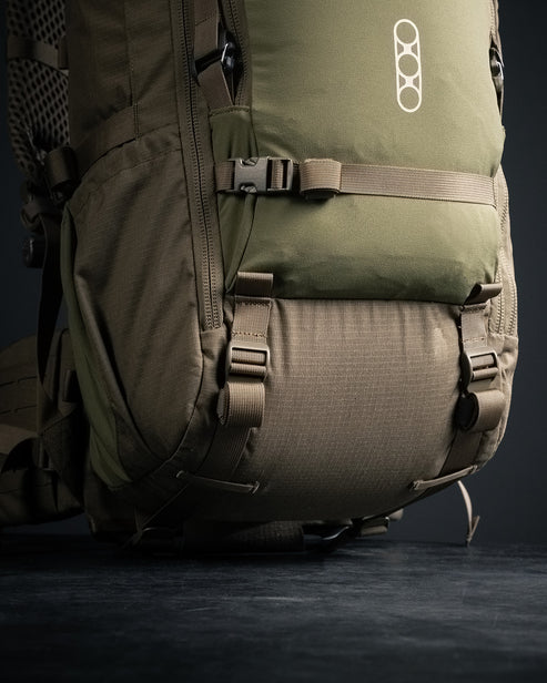 Die Nahaufnahme des EBERLESTOCK Brute 4500 Rucksacks in Grün und Braun zeigt das modulare Design, mehrere Riemen, Schnallen, die Stoffstruktur und das EBERLESTOCK Logo auf der unteren Vorderseite vor einem dunklen Hintergrund.