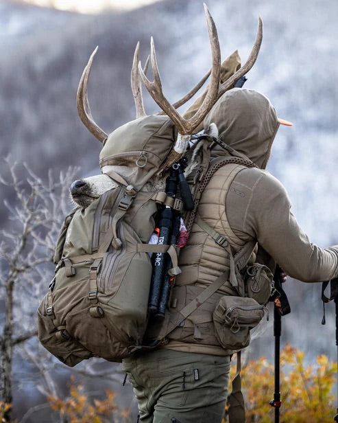 Eine Person in Outdoor-Ausrüstung steht in den Bergen und trägt einen EBERLESTOCK Brute 4500 Rucksack mit Wanderstöcken, Ausrüstung und einem Hirschkopf mit Geweih darauf.