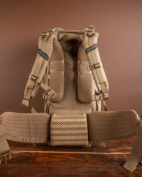 Der modulare Rucksack EBERLESTOCK Brute 3500 wird von hinten gezeigt. Hervorzuheben sind die gepolsterten Mesh-Schultergurte, das belüftete Mesh-Rückenteil und der breite Mesh-Hüftgurt. Der Hintergrund besteht aus einer braunen Wand und einer Holzoberfläche.