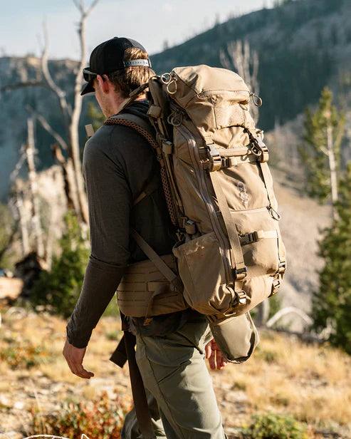 Ein Wanderer in Outdoor-Ausrüstung erkundet einen Bergpfad mit dem EBERLESTOCK Brute 3500 Rucksack. Tannenbäume und ferne Gipfel bilden eine atemberaubende Szene für ein Outdoor-Abenteuer.