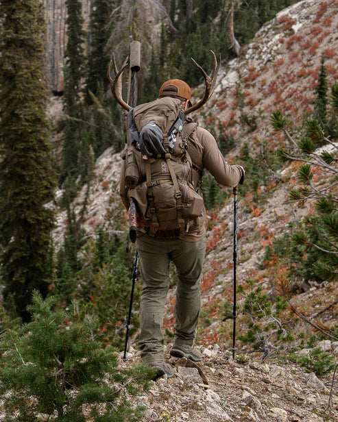 Ein Wanderer auf einer Outdoortour benutzt Trekkingstöcke, während er einen EBERLESTOCK Brute 3500 Rucksack mit aufgesetztem Geweih trägt und einen felsigen Pfad in einem bewaldeten Berggebiet hinaufgeht.