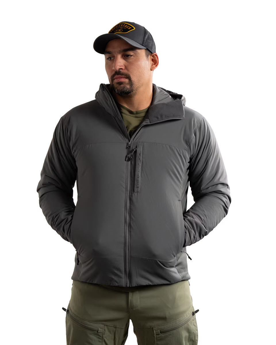 Ein Mann, der eine EBERLESTOCK Alturas Hooded Jacket in Gunmetal, eine grüne Cargohose und eine schwarze Baseballkappe trägt, steht mit den Händen in den Taschen vor einem schlichten weißen Hintergrund.