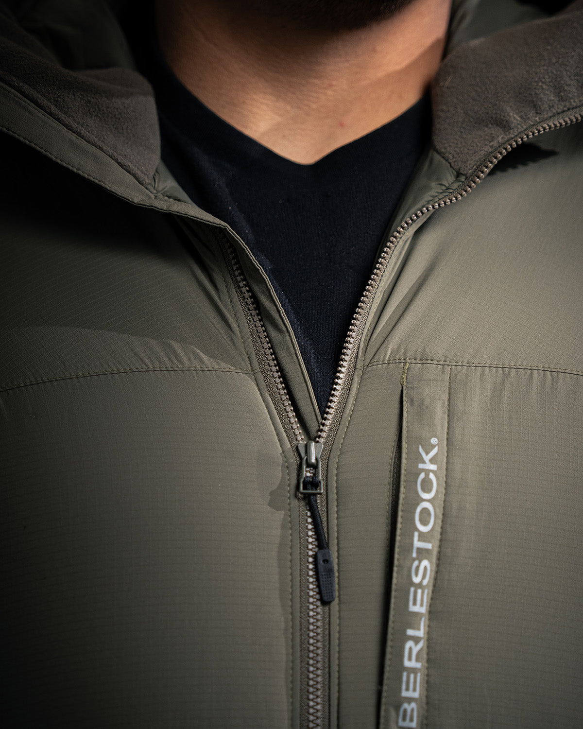 Nahaufnahme einer Person, die die EBERLESTOCK Alturas Hooded Jacket - Gunmetal trägt. Der Reißverschluss ist teilweise geöffnet und gibt den Blick auf ein schwarzes Hemd frei. Das EBERLESTOCK Logo ist auf der Jacke neben dem Reißverschluss zu sehen.