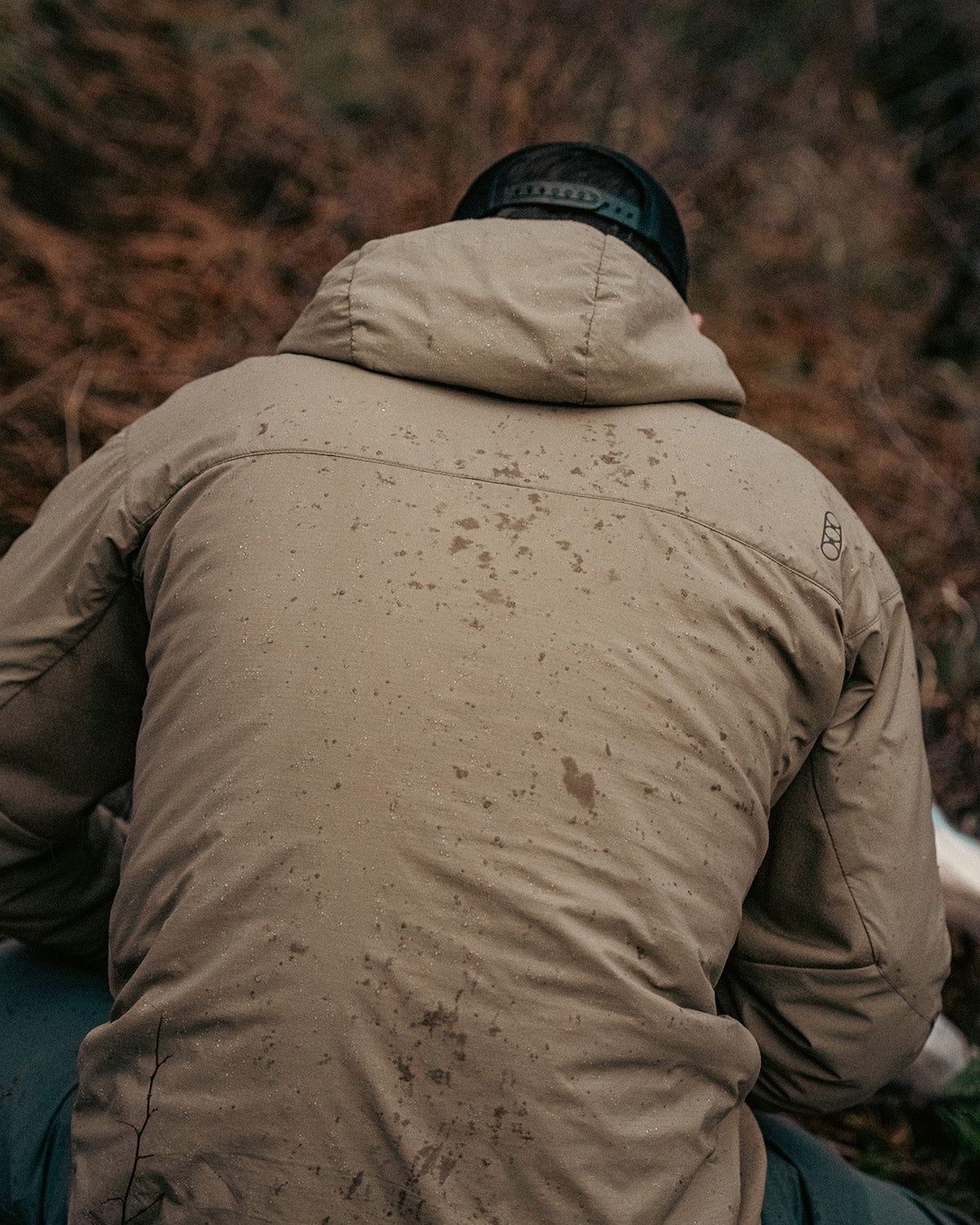 Eine Person in einer EBERLESTOCK Alturas Hooded Jacket - Gunmetal und einer dunklen Mütze sitzt im Freien auf dem Boden, Regentropfen sind auf dem Rücken der Jacke sichtbar. Der verschwommene Hintergrund zeigt erdige Töne.